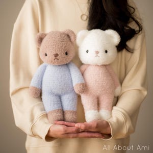 May include: Two crocheted teddy bears, one brown with blue clothing and the other white with pink clothing, are held in the hands of a person wearing a beige sweater.