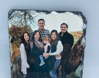 May include: A family portrait printed on a black slate coaster. The family is standing in a wooded area with trees in the background. There are two adults, two children, and a baby in the photo.