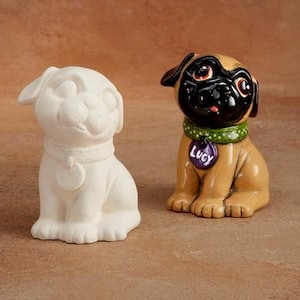 May include: A ceramic pug dog figurine with a black face and brown body. The figurine is sitting with its legs crossed and has a green collar with a purple tag that says "Lucy". There is also a plain white ceramic pug figurine next to it.