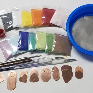 May include: A collection of craft supplies including a bottle, colorful powders in small bags, a blue sieve, various tools, and copper-colored metal shapes. The powders are in shades of yellow, orange, pink, blue, green, and brown.