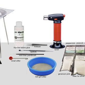 Op de afbeelding: Een set glasfusing-benodigdheden, waaronder een statief en een scherm, een brander, een fles Klyr-Fire-fusers, een kleine zeef, een zeef van 320 grit, pipetten, pincetten, een spatel, een mini-liner-penseel en 10 zakjes gekleurde glasfrit in verschillende tinten blauw, groen, roze, geel, paars en oranje.