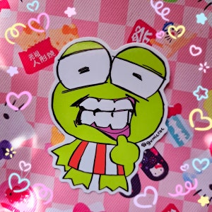 May include: A die-cut sticker featuring a cartoon frog with a wide grin, large white eyes, and a red and white striped shirt. The sticker has the text "@gumicroc" and is set against a pink checkered background with heart and star decorations.