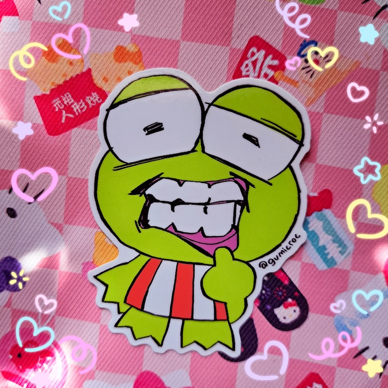 May include: A die-cut sticker featuring a cartoon frog with a wide grin, large white eyes, and a red and white striped shirt. The sticker has the text "@gumicroc" and is set against a pink checkered background with heart and star decorations.
