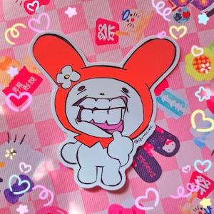 May include: A white sticker featuring a cartoon bunny character with an orange hood and a large, toothy grin. The sticker has a hand-drawn style and is set against a pink checkered background with heart and star-shaped decorations.