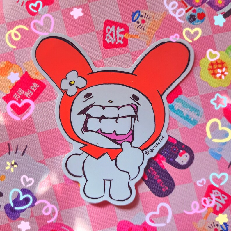 May include: A white sticker featuring a cartoon bunny character with an orange hood and a large, toothy grin. The sticker has a hand-drawn style and is set against a pink checkered background with heart and star-shaped decorations.