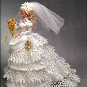 Puede incluir: Muñeca con un vestido de novia blanco de ganchillo hecho a mano con una larga cola y velo. La muñeca sostiene un ramo de flores amarillas con cintas. El vestido presenta intrincados detalles de encaje y una falda escalonada.