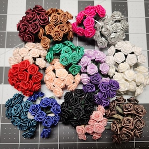 May include: A collection of small, artificial rose bouquets in various colors, including red, pink, silver, white, green, purple, and black. The roses are made of a satin-like material and are arranged in small clusters.