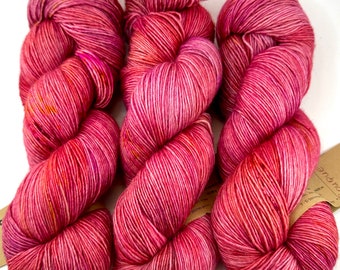 Può includere: Tre matasse di filato tinto a mano nei toni del rosa, del rosso e del viola. Il filato è avvolto strettamente e presenta un motivo cromatico variegato. Le matasse sono disposte fianco a fianco, con un'etichetta visibile sul lato destro.