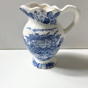 May include: A small, white ceramic pitcher with a blue and white floral pattern. The pitcher has a scalloped rim and a handle. The pattern features a building and trees.