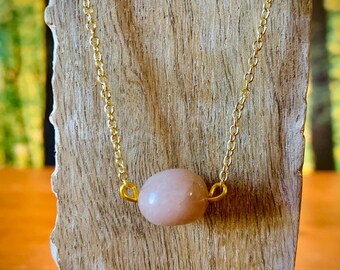 May include: A delicate gold chain necklace with a single, light pink oval gemstone pendant.