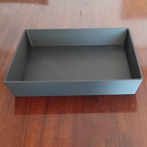 Puede incluir: Una bandeja rectangular negra con un fondo texturizado. La bandeja está hecha de plástico duro.