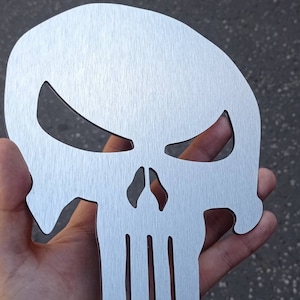 May include: A silver metal skull with a brushed finish. The skull has a menacing expression with a prominent jawline and a series of vertical lines on the lower jaw.