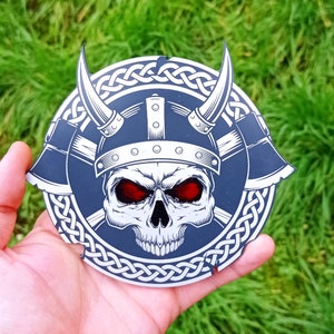 May include: A black and white skull wearing a helmet with a Celtic knot design around the edges. The skull has red eyes and is wearing a helmet with a Celtic knot design around the edges.