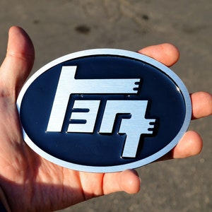 May include: A silver metal emblem with the Toyota logo on a black background. The logo is a stylized "T" with three horizontal lines.