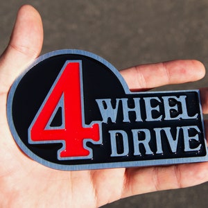 Könnte beinhalten: Ein silbernes Metallemblem mit schwarzem Hintergrund und einer roten Zahl "4" in einem Kreis. Der Text "WHEEL DRIVE" befindet sich unter dem Kreis in silbernen Buchstaben.