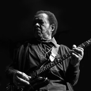 Puede incluir: Una fotografía en blanco y negro de un hombre tocando una guitarra eléctrica. Lleva una camisa negra de botones y una chaqueta negra. La guitarra tiene un cuerpo negro y una placa de protección plateada.