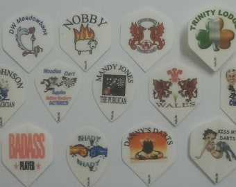 May include: A collection of 12 dart flights with various designs. The designs include a cartoon roadrunner, a sheep on fire, a dragon, a shamrock, a cartoon man, a Welsh dragon, a cartoon woman, a cartoon man with a mustache, a cartoon woman with a red dress, a cartoon man with a fist, a cartoon man with a blue shirt, and a cartoon woman with a red dress. The flights are all white with a black border.