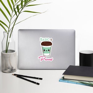 Puede incluir: Una pegatina con una taza de café de dibujos animados con una cara sonriente. La taza es de color verde claro con una tapa marrón y las palabras "BASIC & Proud" en una fuente juguetona. La pegatina está en un portátil.