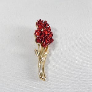 Op de afbeelding: Een goudkleurige metalen broche in de vorm van een boeket bloemen. De bloemen zijn dieprood en de stelen en bladeren zijn goudkleurig. De broche staat tegen een witte achtergrond.