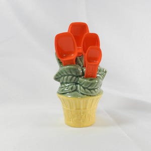 May include: A ceramic measuring spoon set in a flower pot design. The set includes orange spoons nestled in a green leaf arrangement, with a yellow pot base. A decorative and functional kitchen item.