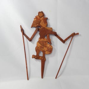 Puede incluir: Una marioneta de sombra de madera marrón con intrincados detalles y patrones tallados. La figura tiene brazos articulados unidos a dos varillas de control de madera delgadas, con una estaca vertical central en la base para soporte.