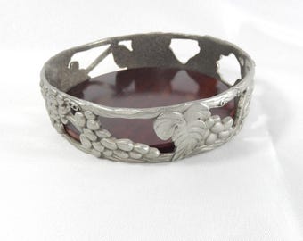 May include: A pewter-colored, circular dish with a dark red interior. The rim is decorated with a grape vine design. The dish is likely used for decorative purposes or to hold small items. The design is detailed and elegant.