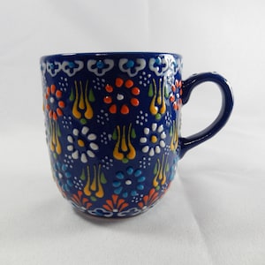May include: A deep blue ceramic mug featuring a raised, multi-coloured floral and tulip pattern. The design includes vibrant orange, yellow, white, and light blue dots and shapes arranged in a repeating motif across the entire exterior surface.
