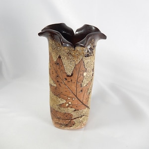 May include: A cylindrical ceramic vase with a ruffled, dark brown glazed rim. The body features a textured, sandy beige finish decorated with embossed brown maple leaf patterns that wrap around the exterior surface of the vessel.