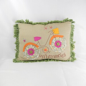 May include: A decorative pillow with a beige background and a stitched bicycle design. The bicycle has pink and orange accents, with floral patterned wheels and the word "Memories" embroidered below. The pillow has green fringe.