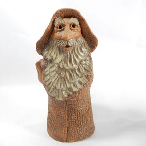May include: A brown clay figurine of a bearded figure wearing a wide-brimmed hat. The figure has a textured coat, a long light-coloured beard, and a moustache. The sculpture is rendered in a monochromatic brown tone with lighter highlights on the beard.