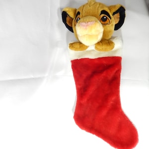 May include: A plush Christmas stocking featuring a Simba lion cub character. The stocking is red with a white cuff, and the lion's head peeks out from the top. The lion has tan fur, black-tipped ears, and a pink nose.