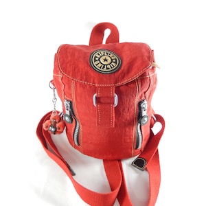 Peut inclure: Un petit sac à dos rouge avec une poignée supérieure et des bretelles réglables. Le sac à dos comporte un compartiment principal zippé, un rabat avant et un petit écusson rond. Le sac a un petit porte-clés singe attaché à la fermeture éclair.
