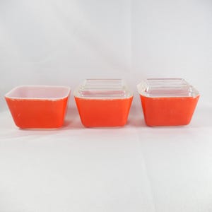 Peut inclure: Trois récipients carrés en verre avec des parois extérieures orange vif et des intérieurs blancs. Deux des récipients sont dotés de couvercles en verre transparent, tandis que le troisième est présenté sans couvercle, révélant son ouverture.