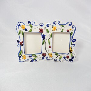 May include: A ceramic double picture frame with a floral design. The frame has two square openings and is decorated with blue, yellow, and red flowers and green leaves. The frame has a white background and a blue border.