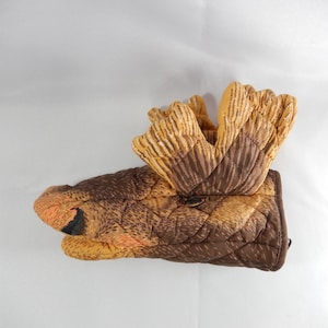 Puede incluir: Un guante de cocina acolchado con forma de cabeza de alce. El guante presenta tonos marrones, beige y naranja, con una nariz negra y un diseño de cuernos detallado. Está diseñado para proteger las manos del calor.