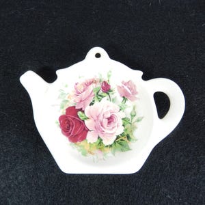 May include: White ceramic teapot-shaped tea bag holder with a floral rose design in pink and red. The holder has a handle and a small hole for hanging.