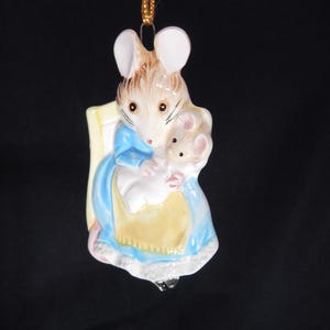 Puede incluir: Adorno de cerámica de una ratona sosteniendo un bebé ratón. La madre ratona lleva un vestido azul y un delantal amarillo. Los ratones tienen pelaje marrón y blanco. El adorno cuelga de un hilo dorado.