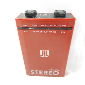 May include: A red AM/FM stereo radio with black volume and tuning knobs. The radio displays AM and FM frequency bands, and the word "STEREO" is printed on the front. The brand name "Jil" is printed on the front of the radio.