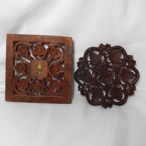 May include: Two wooden decorative pieces. One is square with floral cutouts and a central design. The other is round with a leaf pattern. Both are brown and appear to be hand-carved.