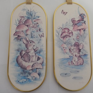May include: Two oval-shaped wall decor pieces with a gold frame. The left piece features a brown rabbit sitting in a mushroom with other mushrooms and a butterfly in the background. The right piece features a brown mouse standing in a pond with lily pads and a butterfly in the background.