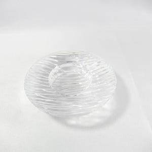 Peut inclure: Un bougeoir en verre transparent de forme circulaire avec une surface texturée à motifs ondulés. Le centre présente une zone circulaire en creux pour une bougie. Le verre est transparent et reflète la lumière, révélant un logo de marque gravé.