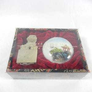 May include: A rectangular gift box with a clear lid containing a beige stone seal with a carved lion figure and a round white ceramic ink paste container featuring a hand-painted landscape scene with a pagoda, trees, and rocks on red velvet.
