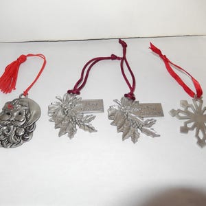 Può includere: Quattro ornamenti natalizi in peltro con nastri rossi e bordeaux. Un ornamento presenta un viso di Babbo Natale, un altro ha un ramo di agrifoglio con il testo "Buon Natale", e il terzo ornamento ha un ramo di agrifoglio con il testo "Buone Feste". Il quarto ornamento è un fiocco di neve.