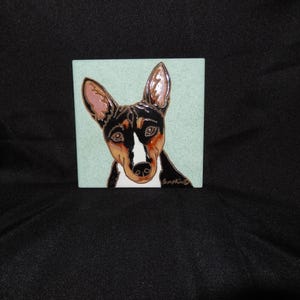 May include: Square ceramic tile with a Basenji dog portrait. The dog has a black and brown coat, a white patch on its face, and large, upright ears. The background is a light blue-green colour.