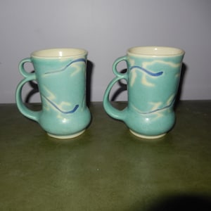 Può includere: Due tazze in ceramica turchese con disegni astratti bianchi e blu. Le tazze hanno un'unica doppia maniglia.