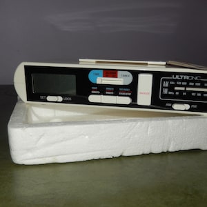 May include: A white and black digital clock radio with a black display and a silver dial. The radio has buttons for setting the time, alarm, and timer. The radio also has buttons for AM and FM radio.