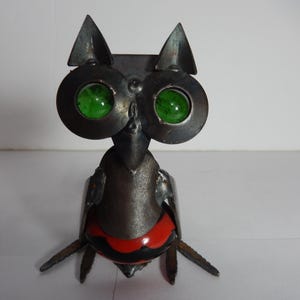 以下が含まれることがあります： 緑色のガラスの目をした金属製のフクロウの彫刻。赤と黒の塗装が施された台座が付いている。