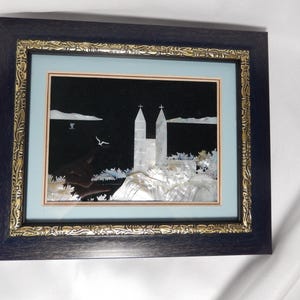 Könnte beinhalten: Gerahmtes Kunstwerk mit einer Kirchenszene, hergestellt aus Perlmutt. Das Kunstwerk ist vor einem schwarzen Hintergrund mit einem hellblauen Rand und in einem dunkelblauen Rahmen mit goldenen Akzenten eingefasst. Das Bild zeigt eine Kirche mit zwei Türmen.
