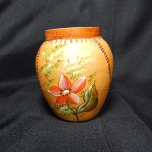 Può includere: Un piccolo vaso in ceramica dipinto a mano con un motivo floreale. Il vaso è di colore arancione chiaro con un bordo arancione più scuro e un motivo cucito sui lati. Un fiore rosso e bianco è dipinto sul davanti.