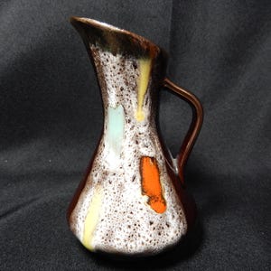 May include: A small, vintage ceramic jug with a brown handle and spout. The body is white with brown speckles and colourful accents of yellow, orange, and light blue. The jug has a unique, textured glaze.